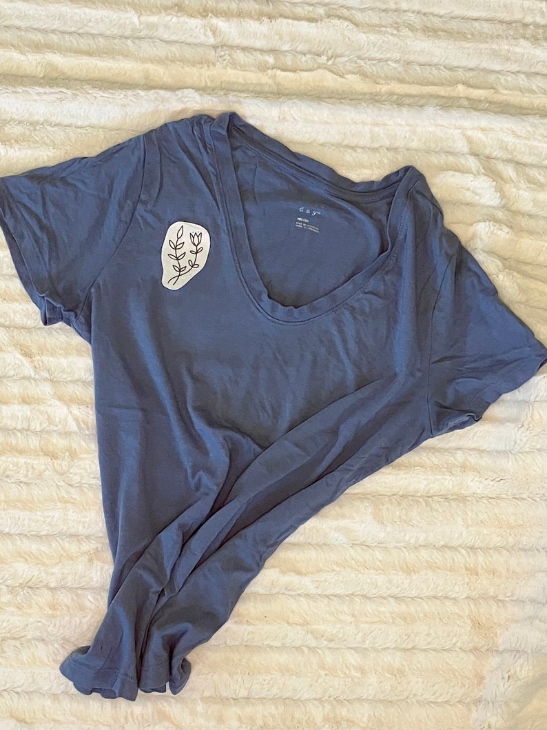Puede incluir: Una camiseta azul con un dise&ntilde;o floral blanco en el lado izquierdo del pecho. El dise&ntilde;o es un simple dibujo de l&iacute;nea de una rama con hojas y flores.