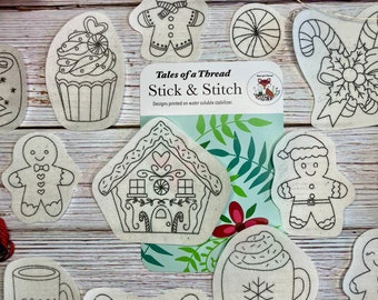 May include: A set of 11 iron-on embroidery designs featuring various Christmas themes, including a gingerbread house, a gingerbread man, a candy cane, a cupcake, a mug of cocoa, and a snowflake. The designs are printed on water-soluble stabilizer, making them easy to use. The text "Tales of a Thread Stick & Stitch" is printed on a card behind the designs.