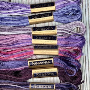 May include: A collection of embroidery floss in shades of purple, lavender, and pink. Each skein is labeled "Seasons" and "Embroidery Six Strand", with the text "100% Cotton Made in Japan". The floss is 8 meters in length.