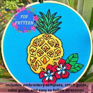 Può includere: Ananas ricamato con foglie verdi e fiori di ibisco rossi su uno sfondo blu. Il ricamo è in un cerchio e il testo "PDF PATTERN" è visibile in alto a sinistra.