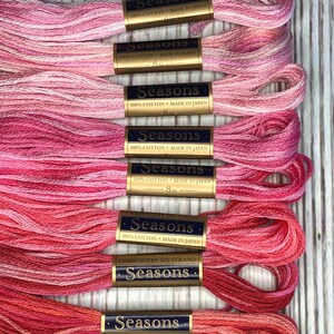 May include: A stack of eight spools of pink, red and white embroidery floss. Each spool is labelled "Seasons" and "100% Cotton Made in Japan".
