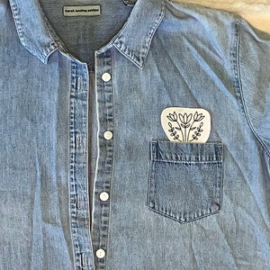 Puede incluir: Una camisa vaquera azul claro con un bolsillo blanco y un dise&ntilde;o floral negro en el bolsillo. La camisa tiene un cierre de botones en la parte delantera y un cuello. La etiqueta de la camisa dice "marsh landing petites".