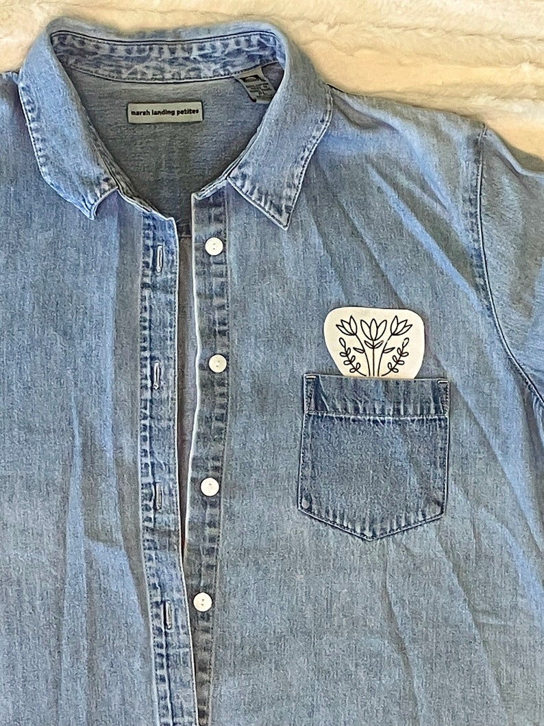 Puede incluir: Una camisa vaquera azul claro con un bolsillo blanco y un dise&ntilde;o floral negro en el bolsillo. La camisa tiene un cierre de botones en la parte delantera y un cuello. La etiqueta de la camisa dice "marsh landing petites".