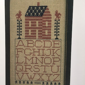May include: A framed cross-stitch sampler with a red, white, and blue house design and the alphabet in red. The sampler is dated 1988.