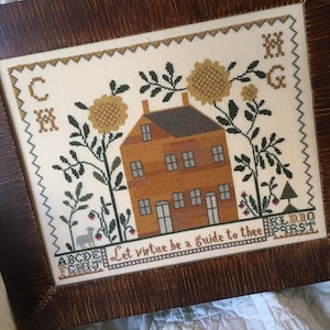 May include: A framed cross-stitch sampler with a brown wooden frame. The sampler features a house with a brown roof and yellow walls, surrounded by green plants and yellow flowers. The text "Let virtue be a guide to thee" is stitched below the house.