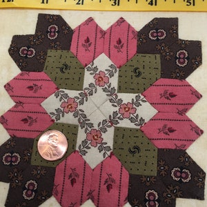 May include: A quilt block with a starburst pattern in shades of brown, pink, and green. The block is made up of smaller pieces of fabric sewn together. A penny is placed in the center of the block for scale.
