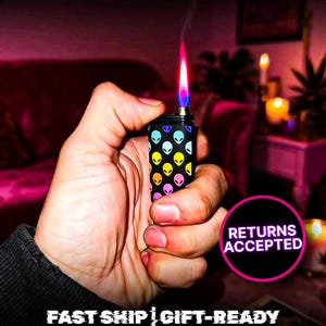 May include: A black lighter with a colorful alien head pattern, held in a hand. The lighter's flame is a vibrant mix of red, orange, and blue. Text on the image reads "OUT-OF-THIS-WORLD DESIGN" and "RETURNS ACCEPTED".