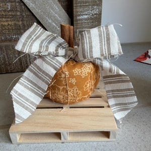 Può includere: Una zucca in tessuto color giallo senape con un motivo floreale bianco, sormontata da un gambo a bastoncino di cannella e un fiocco in nastro sfilacciato a righe beige e bianche, poggiata su una piccola piattaforma a pallet in legno chiaro.