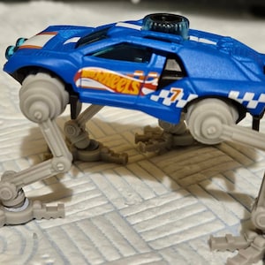 Może przedstawiać: Niebieski plastikowy samochód z białym paskiem i napisem "Hot Wheels" na boku. Samochód ma cztery szare nogi i czarny spojler. Samochód stoi na biało-szarej powierzchni.
