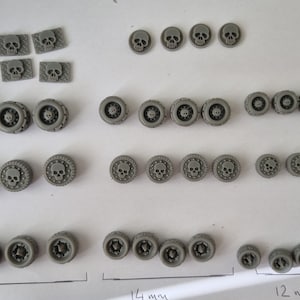 Puede incluir: Una colección de ruedas de plástico gris en forma de calavera de varios tamaños, que van de 12 mm a 14 mm. Las ruedas están dispuestas en filas sobre una superficie blanca.