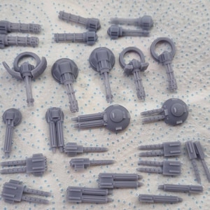 Puede incluir: Una colección de 20 cañones en miniatura de plástico gris, algunos con cañones giratorios, otros con cañones fijos. Los cañones están dispuestos sobre una superficie blanca.