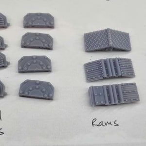 May include: A set of 3D printed model parts for a miniature vehicle. The set includes wheel covers, rams, and track pieces. The parts are grey and have a textured surface.