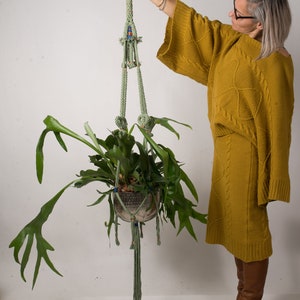 Puede incluir: Un macetero de macramé verde con una maceta de cerámica marrón y una planta verde. El macetero está colgado del techo y la planta está en la maceta. Una persona está sosteniendo el macetero en su mano.