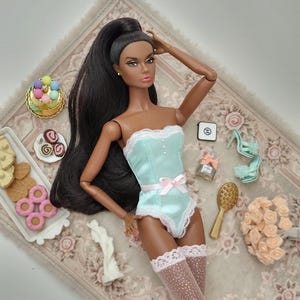 May include: A fashion doll with long black hair, wearing a light blue bodysuit with a pink bow and lace trim, and patterned thigh-high stockings. Accessories include miniature desserts, perfume, a hairbrush, and a bouquet of flowers.