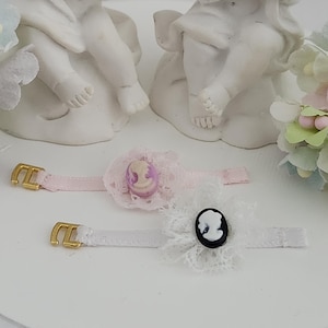 May include: Two decorative hair accessories with cameo designs. One is pink with a cameo in shades of purple and white, the other is white with a black and white cameo. Both have gold-coloured buckles and lace accents.