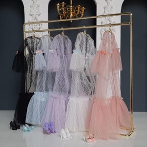 May include: A gold clothing rack with five tulle dresses hanging on it. The dresses are in black, light blue, lavender, white, and pink. There are five pairs of shoes on the floor in front of the rack.