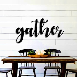 May include: Black metal wall decor with the word "gather" in a cursive font. The decor is hanging above a wooden dining table with four black chairs.