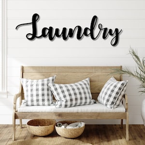 May include: Black cursive "Laundry" sign mounted on a white shiplap wall above a wooden bench. The bench has three gray and white checkered pillows, two woven baskets, and a neutral rug.