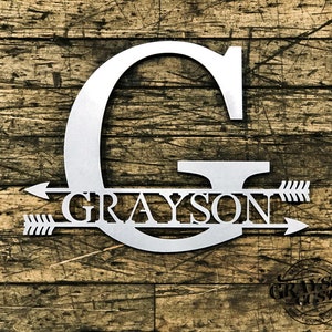 May include: A metal sign with the letter G and the name Grayson with arrows on either side. The sign is made of metal and has a silver finish.