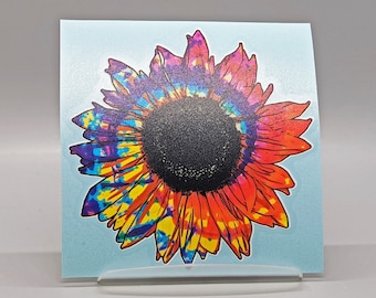 May include: A tie-dye sunflower sticker with a blue, purple, yellow, orange, and red color scheme. The sunflower is on a light blue background.