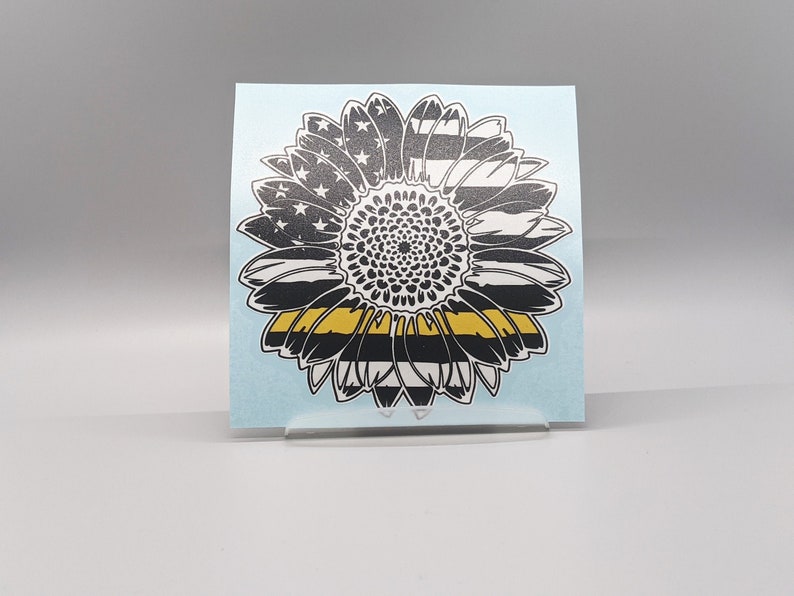 May include: Black and white sunflower with a yellow and black stripe across the center. The sunflower is made to look like an American flag with a thin yellow stripe across the center.