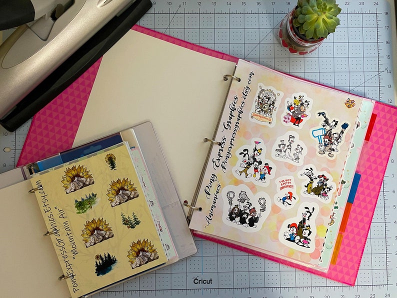 May include: A pink binder with a white interior and a clear plastic sleeve containing stickers. The stickers feature a mountain range with a sunflower and a forest scene. The binder also contains a page of colorful cartoon stickers with the text "Pony Express Graphics -  Ponyexpressgraphics.etsy.com".