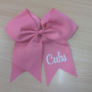 May include: A large pink hair bow made of grosgrain ribbon with a central knot. The bow features two long, pointed tails, and the word "Cubs" is printed in white script lettering on the right-hand tail of the accessory.