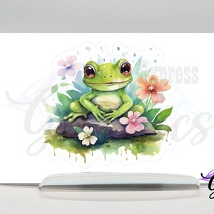 May include: A watercolor illustration of a green frog sitting on a rock surrounded by pink, white, and orange flowers. The frog has large, brown eyes and a wide smile.