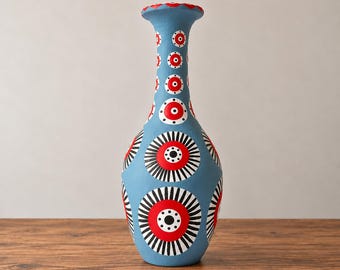 Może przedstawiać: Wysoki, smukły wazon ceramiczny o matowym, jasnoniebieskim kolorze podstawowym. Powierzchnia ozdobiona jest powtarzającym się wzorem dużych okrągłych motywów z jaskrawoczerwonymi środkami, czarnymi promienistymi liniami i małymi białymi kropkami.