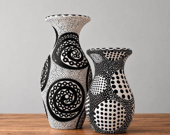Może przedstawiać: Dwa ceramiczne wazony z czarno-białym wzorem w kropki. Wyższy wazon ma rozszerzany kołnierz i spiralne wzory, podczas gdy krótszy, szerszy wazon prezentuje wzór z czarnych i białych kół, oba ozdobione drobnym kropkowaniem.