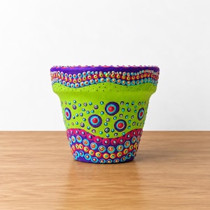 May include: A terracotta plant pot decorated with hand-painted dot art in vibrant shades of lime green, purple, magenta, orange, and yellow. The design features concentric circles and wavy bands of dots covering the exterior surface of the pot.