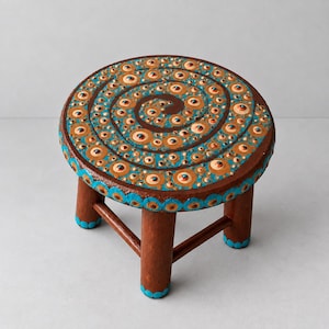 May include: A small, round, brown wooden stool featuring a hand-painted spiral design on the top surface. The pattern consists of concentric circles in shades of light tan, dark brown, and bright turquoise, with matching turquoise dots on the legs.