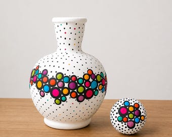 Może przedstawiać: Biała ceramiczna karafka z wąską szyjką i pasującym kulistym korkiem. Powierzchnia ozdobiona jest wzorem z małych czarnych kropek oraz centralnym poziomym pasem większych, jaskrawych, wielokolorowych kół w odcieniach różu, błękitu, pomarańczu i zieleni.