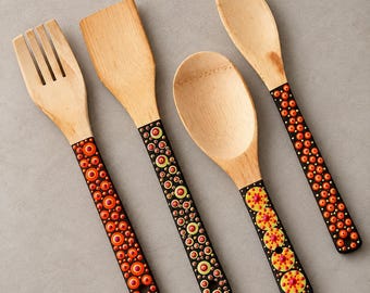 Może przedstawiać: Zestaw czterech drewnianych przyborów kuchennych, w tym widelca, łopatki i dwóch łyżek. Każdy uchwyt jest pomalowany na czarno i ozdobiony wzorem małych, żywych pomarańczowych, żółtych i czerwonych kropek ułożonych w koliste wzory.
