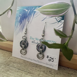 May include: Silver-toned earrings with a chainmail design. Each earring features a small, dark-colored ring connected to a larger, silver ring, with smaller rings dangling below. The earrings are displayed on a card with the text "The Steel Sparrow Handmade Jewelry".
