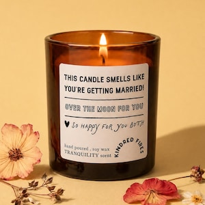 May include: A lit soy wax candle in a dark amber glass jar with a white label reading THIS CANDLE SMELLS LIKE YOU'RE GETTING MARRIED! OVER THE MOON FOR YOU SO HAPPY FOR YOU BOTH. The candle is surrounded by several dried pink and white flowers.