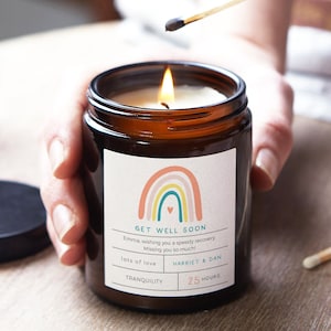 May include: A brown glass candle jar with a white label. The label features a rainbow design with a heart at the bottom. The text on the label reads "Get Well Soon", "Emma wishing you a speedy recovery. Missing you so much!"  "lots of love", "HARRIET & DAN", "TRANQUILITY", and "25 HOURS".