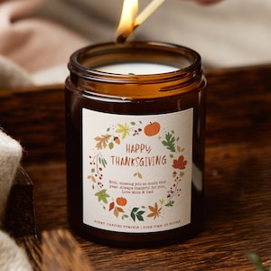 May include: A brown glass candle with a white label that says "Happy Thanksgiving" and features a wreath of autumn leaves, acorns, and pumpkins. The text on the label reads "Erin, missing you so much this year. Always thankful for you. Love Mom & Dad." The candle is scented with candied pumpkin and has a burn time of 25 hours.