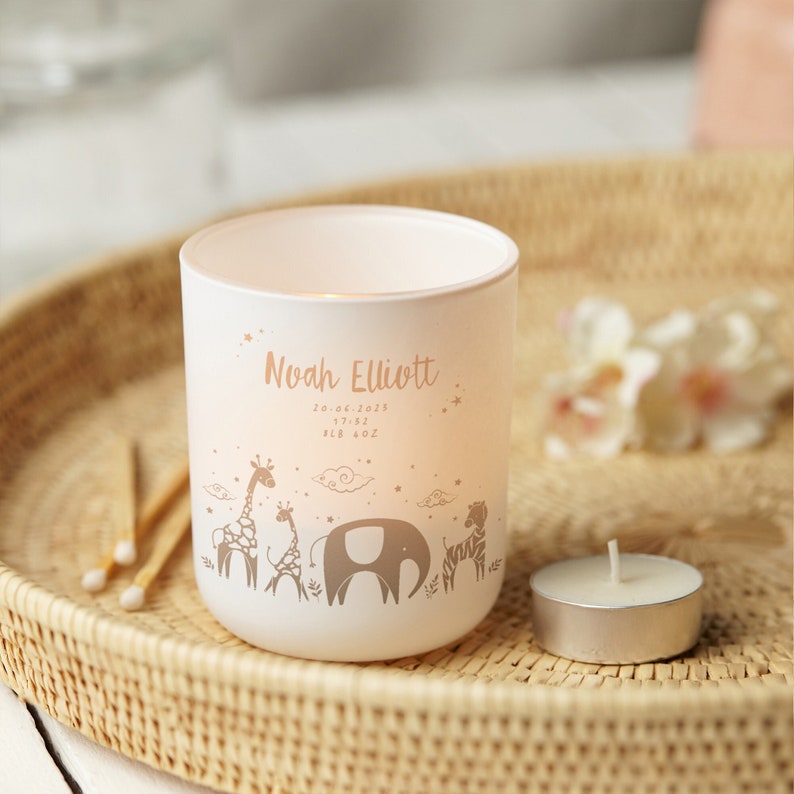 Puede incluir: Un portavelas blanco con un dise&ntilde;o decorativo de animales de safari, incluyendo jirafas, un elefante y una cebra. El portavelas tiene el nombre "Noah Elliott" y detalles de nacimiento. Una vela de t&eacute; encendida est&aacute; cerca.