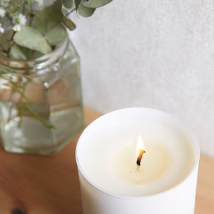 May include: A white candle with a single wick burning in a white jar. The candle is on a wooden surface.