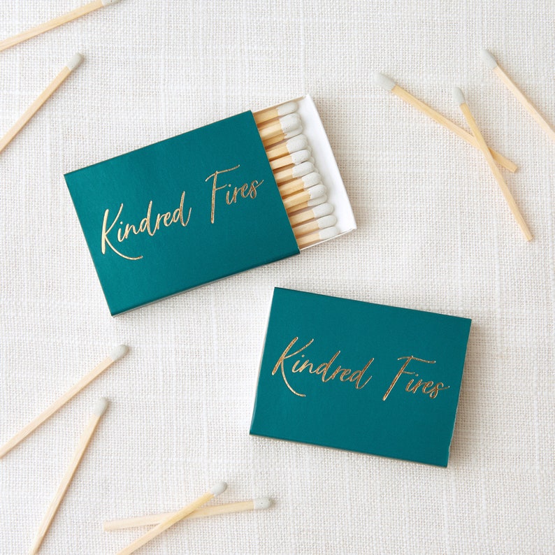 May include: Two teal blue matchboxes with gold lettering that reads "Kindred Fires". The matchboxes are on a white textured surface with wooden matches scattered around them.