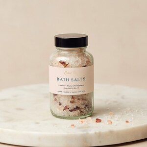 May include: A glass jar of bath salts with a white label that reads "Kanded F" in gold lettering. The label also says "BATH SALTS", "Lavender, Thyme & Ylang Ylang Essential Oil Blend", and "HAND POURED IN SMALL BATCHES".