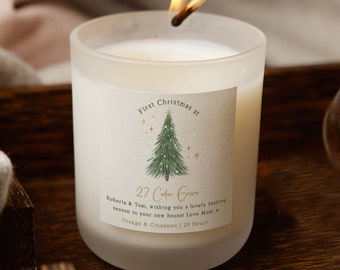 May include: A white frosted glass candle with a label that reads "First Christmas at 27 Cedar Grove Roberta & Tom, wishing you a lovely festive season in your new house! Love Mum x Orange & Cinnamon | 25 Hours". The candle has a green and white Christmas tree illustration with gold stars.