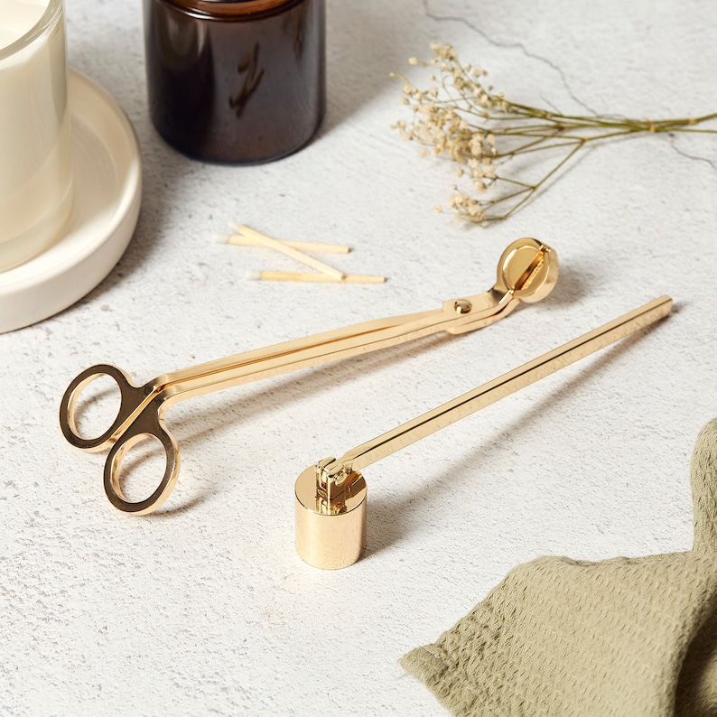 May include: Gold candle wick trimmer and snuffer tools. The trimmer has a pair of scissors and a round snuffer. The snuffer is attached to a long, thin metal rod.