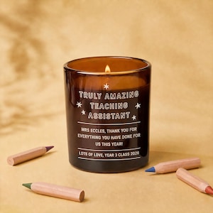 May include: A brown glass candle in a cylindrical jar with a lit wick. The text reads: TRULY AMAZING TEACHING ASSISTANT, MRS ECCLES, THANK YOU FOR EVERYTHING YOU HAVE DONE FOR US THIS YEAR! LOTS OF LOVE, YEAR 3 CLASS 2026. Three coloured pencils lie nearby.
