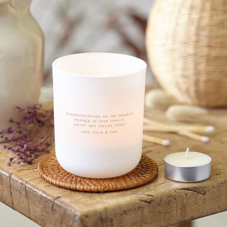 Puede incluir: Un portavelas de vidrio esmerilado blanco con el texto "Felicidades al nuevo miembro de tu familia. &iexcl;Disfruta este momento especial! Love Julia & Tom." Se encuentra sobre un posavasos tejido, con una vela de t&eacute; cerca.