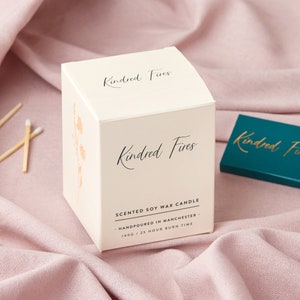 May include: White box with the text "Kindred Fires" on the front and top. The box is a candle container with the text "Scented Soy Wax Candle", "Handpoured in Manchester", and "140g / 25 Hour Burn Time" printed on the front.