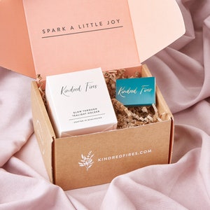 May include: A brown cardboard box with a white lid, containing a white tealight holder with the text "Kindred Fires Glow Through Tealight Holder Crafted in Manchester" and a teal matchbox with the text "Kindred Fires". The box has the text "KINDREDFIRES.COM" printed on the bottom.