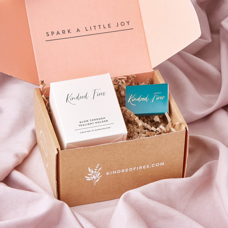 May include: A brown cardboard box with a white lid, containing a white tealight holder with the text "Kindred Fires Glow Through Tealight Holder Crafted in Manchester" and a teal matchbox with the text "Kindred Fires". The box has the text "KINDREDFIRES.COM" printed on the bottom.