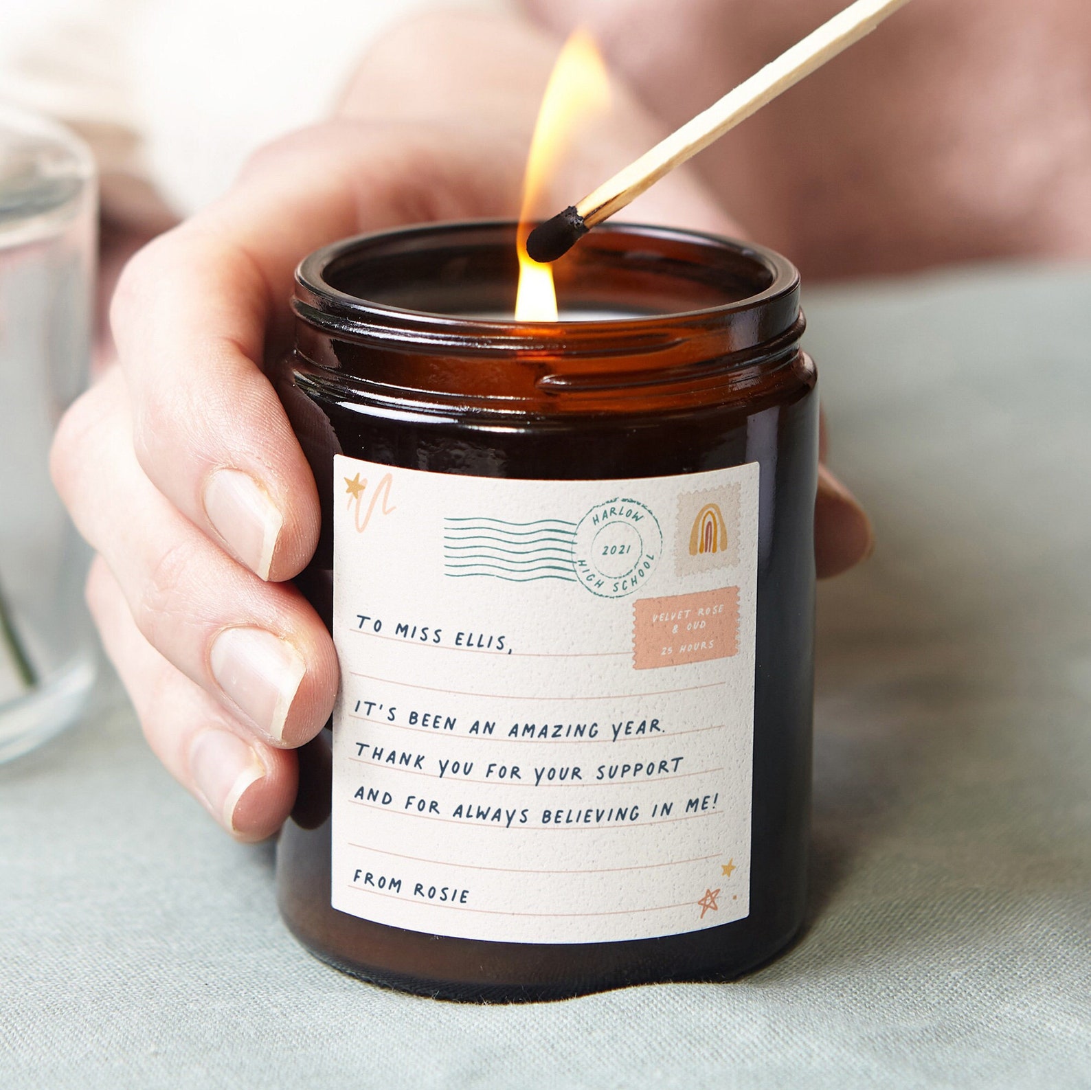 A personalized post-card label on a candle being lit with a match, with one hand holding the candle. The copy on the candle reads "TO MISS ELLIS, IT'S BEEN AN AMAZING YEAR. THANK YOU FOR YOUR SUPPORT AND FOR ALWAYS BELIEVING IN ME. FROM ROSIE" with additioal details like a pink rainbow "stamp", a banner saying the year and highschool (2021, Harlow High School), and a label detailing the scent and burn time (Velvet Rose & Oud, 25 hours)